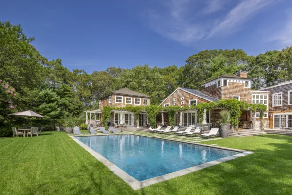 East Hampton retreat poolside with chaise lounge chairs, umbrellas with clapboard house in the background.